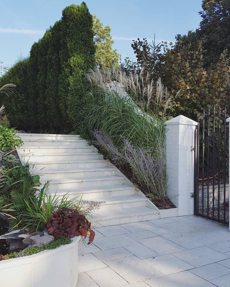 Białe schody z kamienia prowadzące w górę, otoczone wysokimi trawami ozdobnymi i roślinnością, z elementami małej architektury ogrodowej i czarną, kutą bramą w tle.