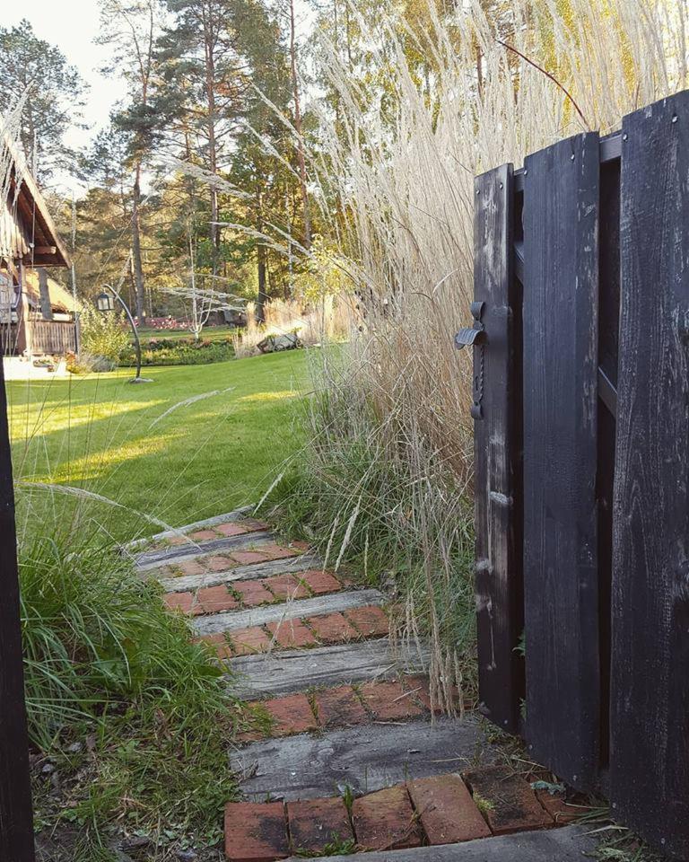 Otwarta, ciemna, drewniana furtka z metalowym uchwytem, prowadząca do ogrodu z ceglaną ścieżką, wysokimi trawami ozdobnymi i domem w tle.