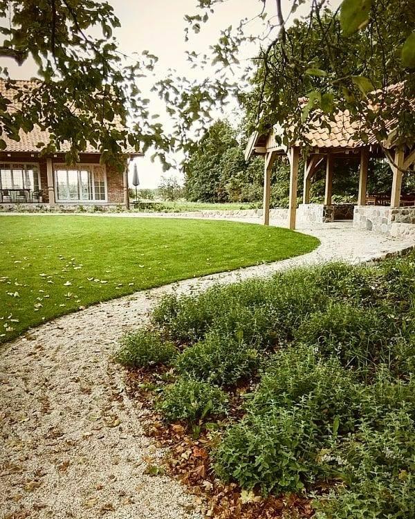 Widok na zadbany ogród z równo przystrzyżonym trawnikiem, kamienną ścieżką i altaną z kamiennymi elementami, w tle dom z cegły.