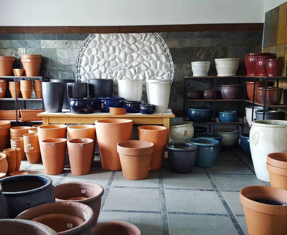 Różnorodność ceramicznych donic ogrodowych w odcieniach terakoty, granatu i bieli, ustawionych na drewnianym stole i metalowych regałach na tle ściany z kamienia łupanego i dekoracyjnej mozaiki.