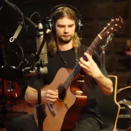 Młody muzyk z długimi włosami gra na gitarze akustycznej w studiu nagraniowym, z mikrofonem pojemnościowym i słuchawkami na uszach, widoczne perkusja w tle.