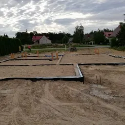 Fundamenty domu jednorodzinnego w trakcie budowy, widoczne szalunki i rury instalacyjne, w tle zarys zabudowań mieszkalnych pod pochmurnym niebem.