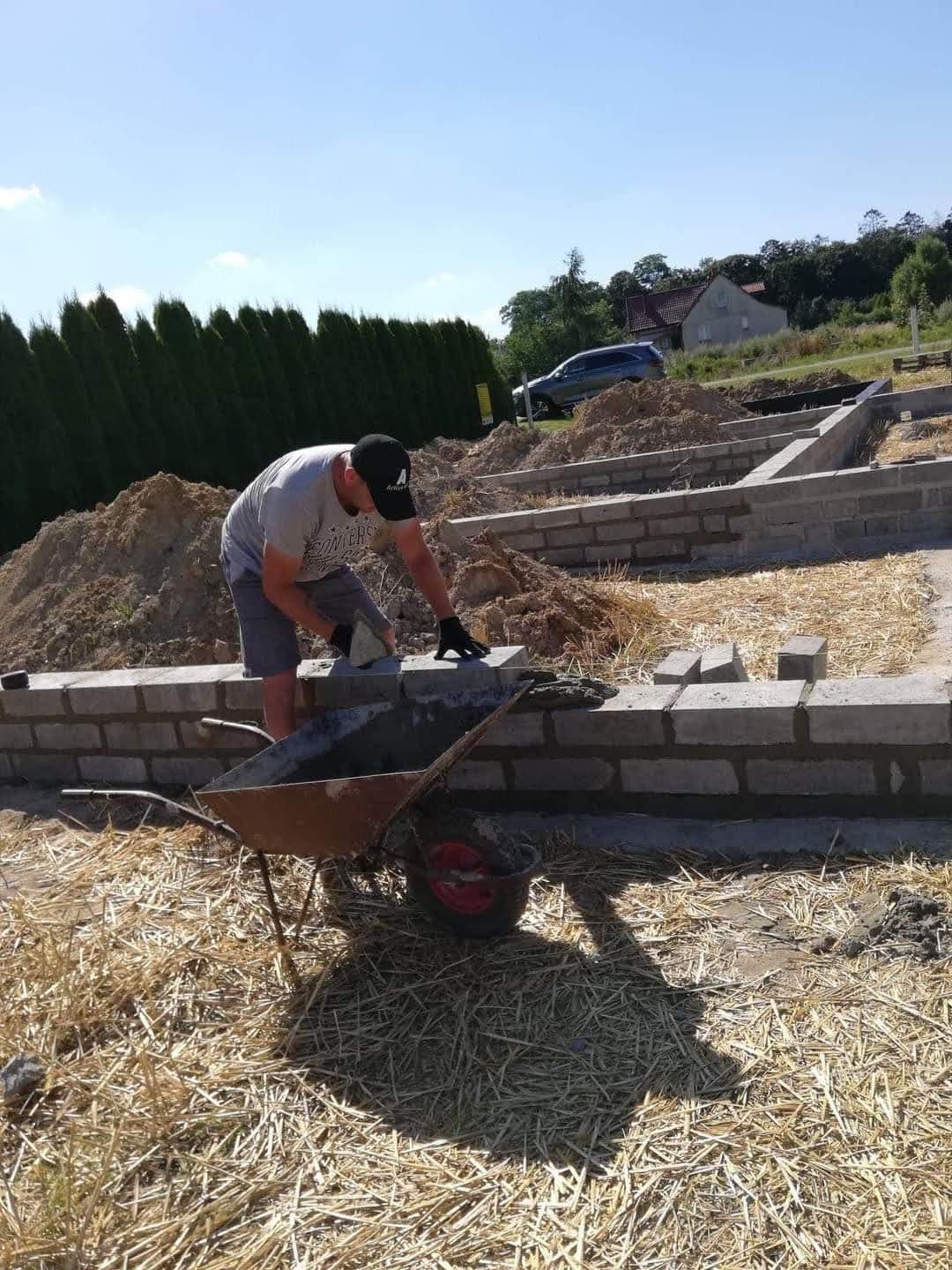 Murarz układa bloczki fundamentowe na zaprawie, widoczna taczka z zaprawą, w tle dom i zielony żywopłot.