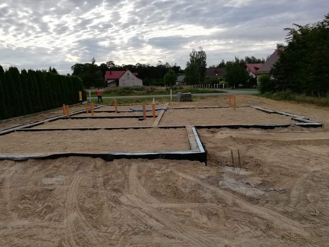 Fundamenty domu jednorodzinnego w trakcie budowy, widoczne szalunki i rury instalacyjne, w tle zarys zabudowań mieszkalnych pod pochmurnym niebem.
