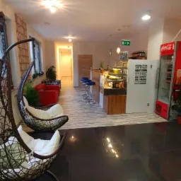 Wnętrze kawiarni lub hotelu SPA z dwoma wiszącymi fotelami rattanowymi w pierwszym planie, barem z ekspozycją ciast i napojów, oraz lodówką Coca-Coli w tle.