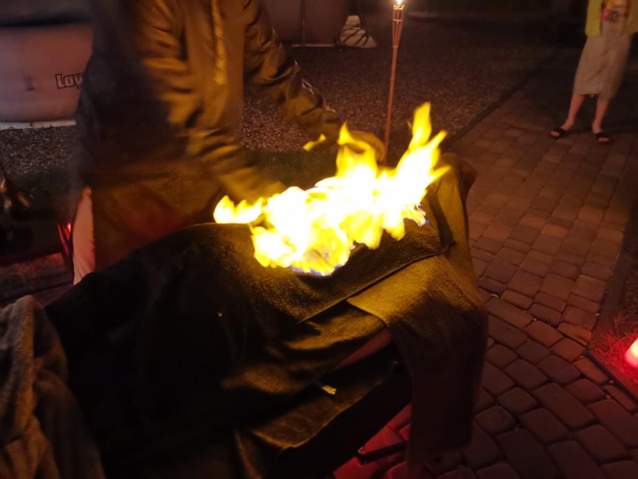 Osoba w kurtce podpala ogień na metalowej konstrukcji podczas wieczornego wydarzenia z pochodniami w tle.