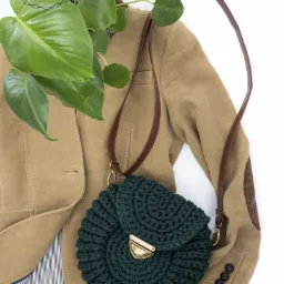 Szmaragdowa, okrągła torebka z klapką na brązowym pasku, leżąca na beżowej marynarce z rośliną w tle.