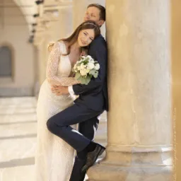 Para młoda w objęciach, oparta o kolumnę w słoneczny dzień. Panna młoda w sukni z długim rękawem, trzyma bukiet białych róż. Pan młody w granatowym garniturze.