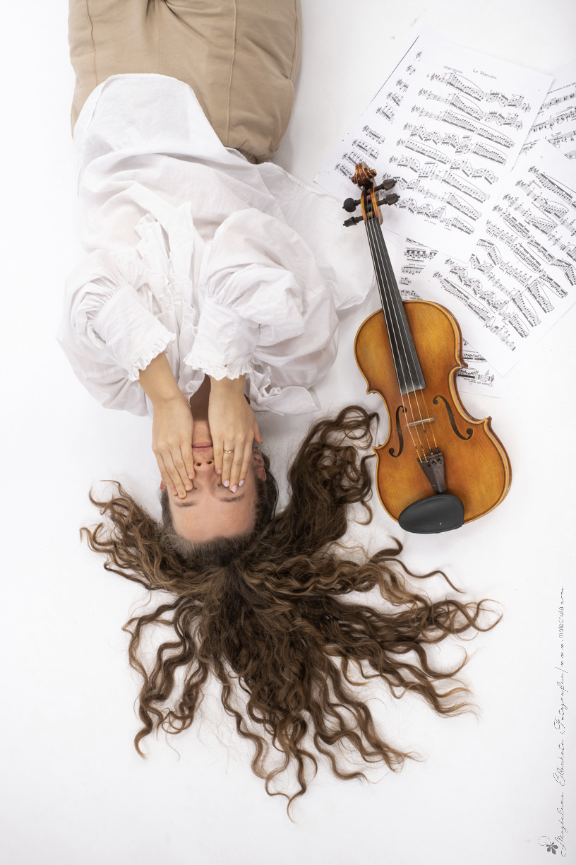 Kobieta w białej sukience leżąca na białym tle, zakrywa oczy dłońmi, obok leżą nuty i skrzypce.