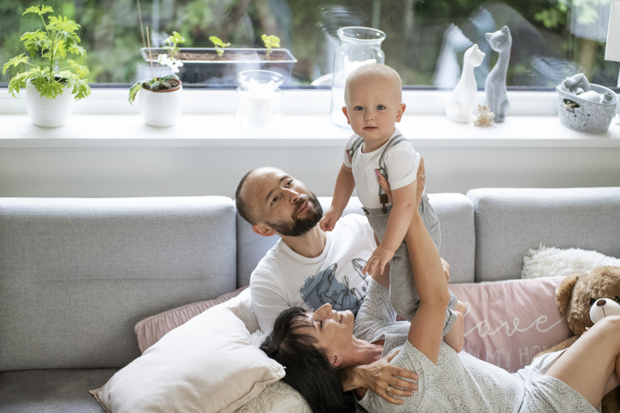 Rodzinne ujęcie: mama leżąc trzyma na rękach synka w szarych szelkach, tata uśmiecha się obok, w tle okno z roślinami i dekoracjami, jasna kanapa i poduszki.