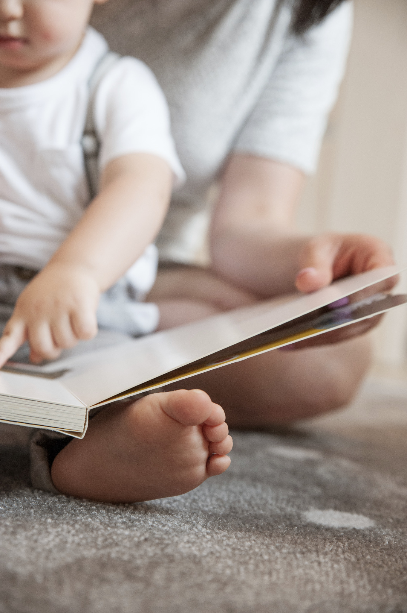 Mama z dzieckiem siedzą na dywanie i oglądają książkę z kolorowymi ilustracjami, z bliska widoczne stopy dziecka.