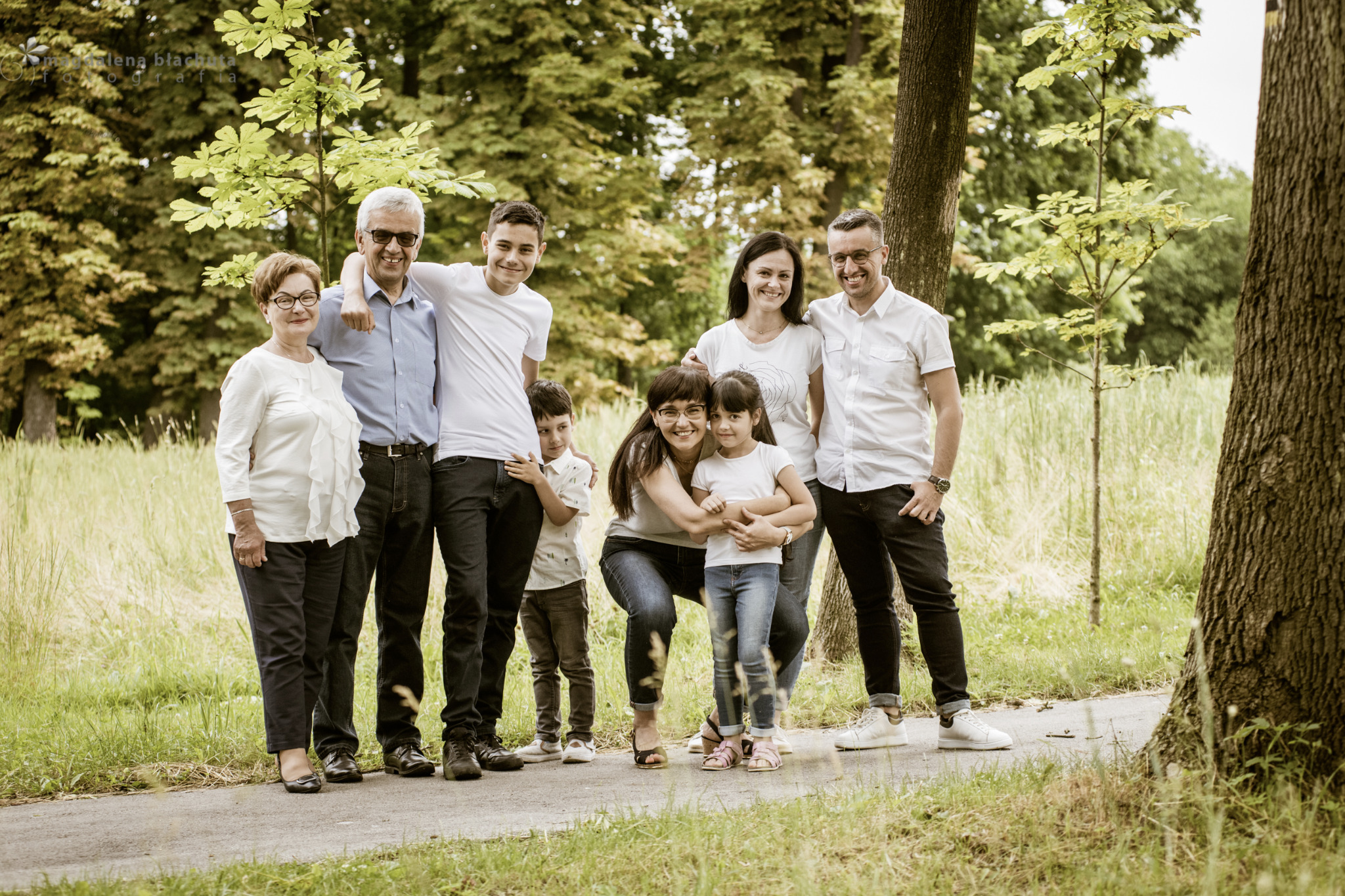 Wielopokoleniowa rodzina: dziadkowie, rodzice i dzieci, pozuje razem na leśnej ścieżce, uśmiechnięci i w jasnych ubraniach.