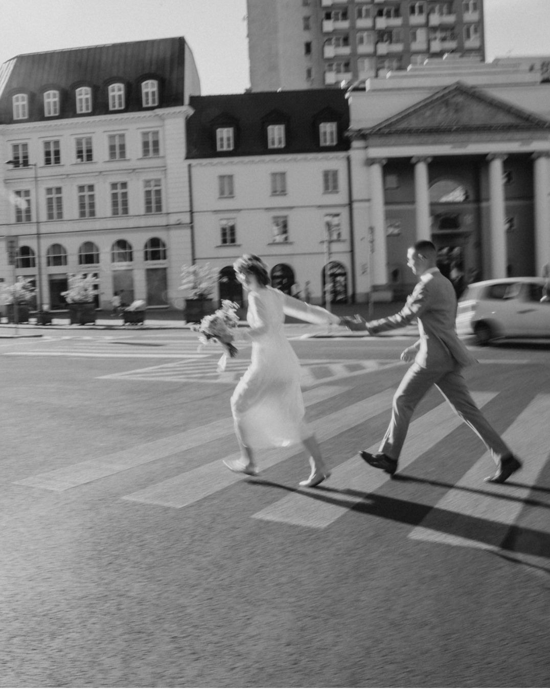Czarno-białe zdjęcie pary młodej biegnącej przez przejście dla pieszych na tle miejskiej architektury. Panna młoda trzyma bukiet, oboje uśmiechnięci, w ruchu.