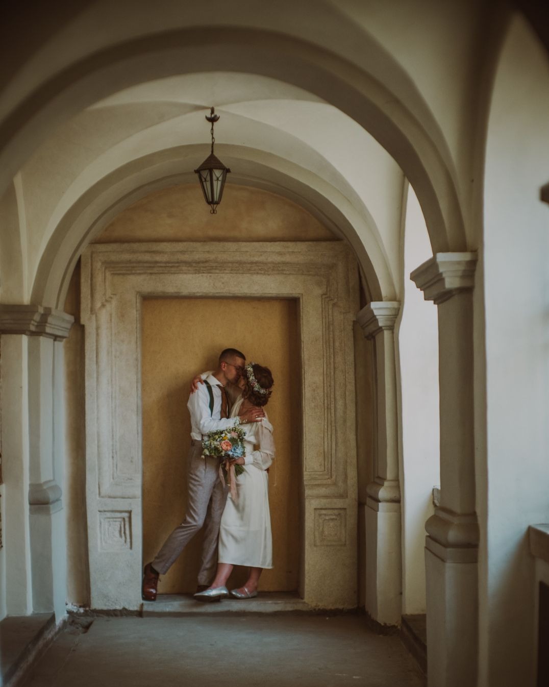 Para młoda w objęciach, pocałunek na tle architektonicznego portalu z łukami. Bukiet w rękach panny młodej, on w okularach i szelkach, ona w wianku.