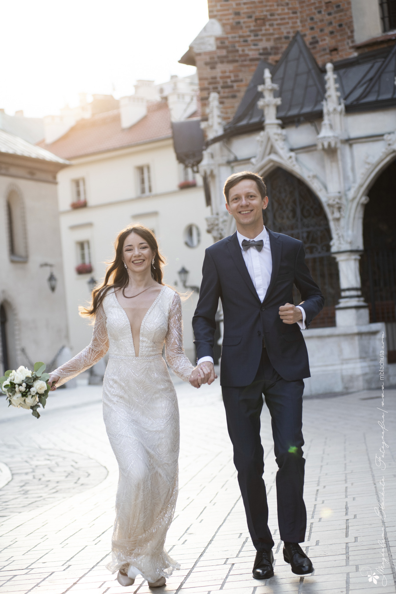 Uśmiechnięta para młoda biegnie trzymając się za ręce na tle zabytkowej architektury Krakowa, panna młoda w sukni z długim rękawem i bukietem białych kwiatów, pan młody w granatowym garniturze...