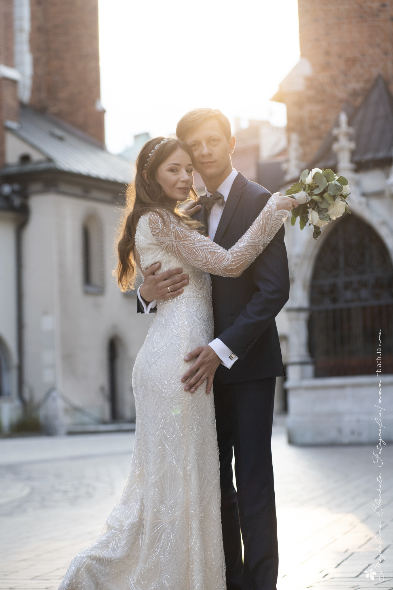 Para młoda w objęciach na tle architektury miejskiej w promieniach zachodzącego słońca; panna młoda w sukni z długim rękawem i wzorem z paciorków, pan młody w granatowym garniturze i muszce, trzyma...