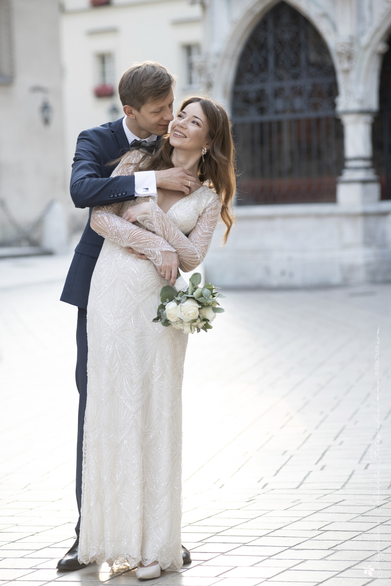 Uśmiechnięta panna młoda w sukni z długim rękawem, trzymająca bukiet białych róż, obejmowana przez pana młodego w granatowym garniturze na tle gotyckiej architektury.