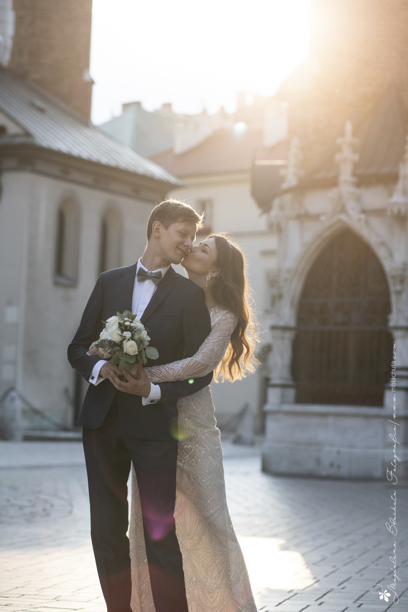 Panna młoda całuje pana młodego trzymającego bukiet białych róż na tle zabytkowej architektury w promieniach słońca.