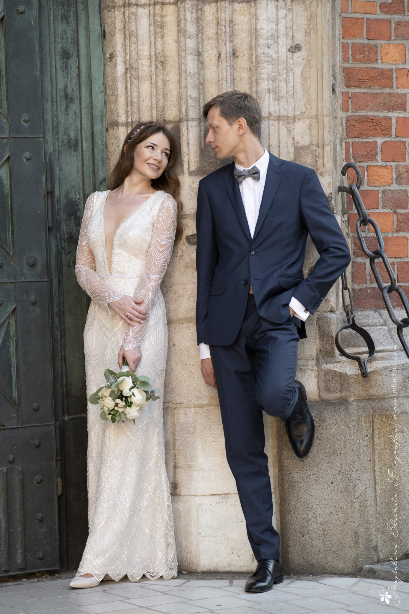 Elegancka para młoda: panna młoda w sukni z długim rękawem i bukietem, pan młody w granatowym garniturze z muchą, pozujący na tle zabytkowej architektury.