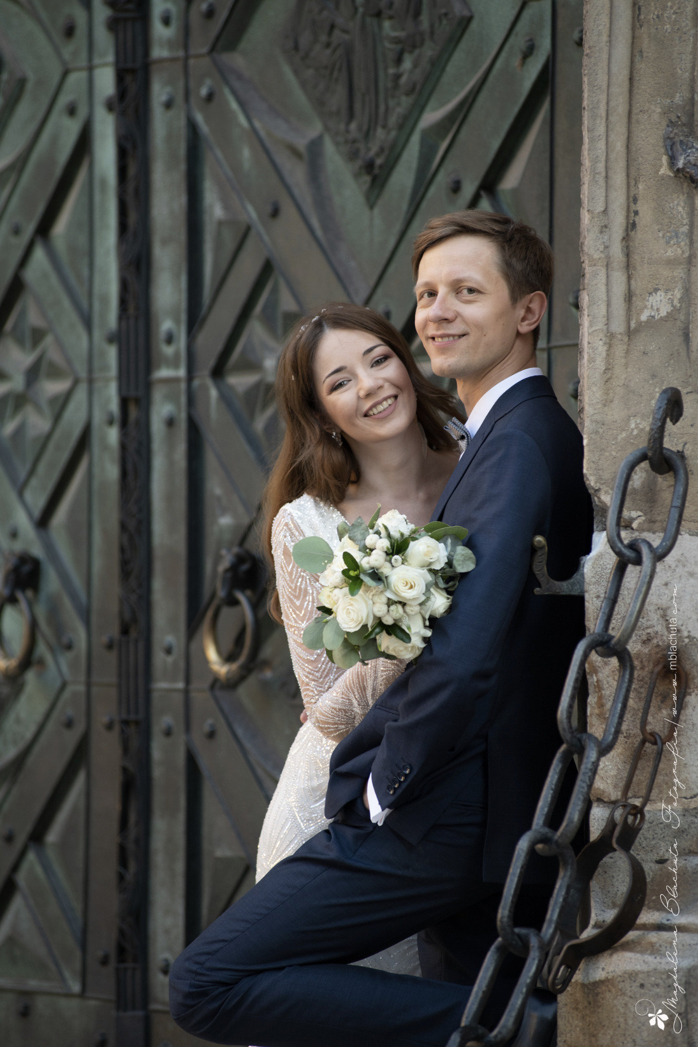 Uśmiechnięta para młoda w objęciach pozuje na tle masywnych, zdobionych drzwi i kamiennego muru z łańcuchami.