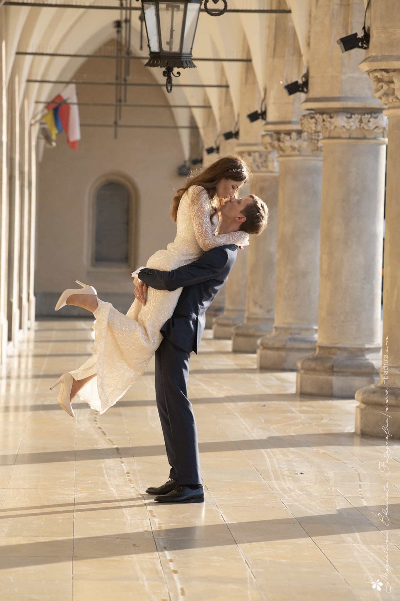 Para młoda: mężczyzna w ciemnym garniturze trzyma na rękach kobietę w długiej, zdobionej sukni ślubnej i szpilkach, całując ją na tle kolumnady z lampionami.