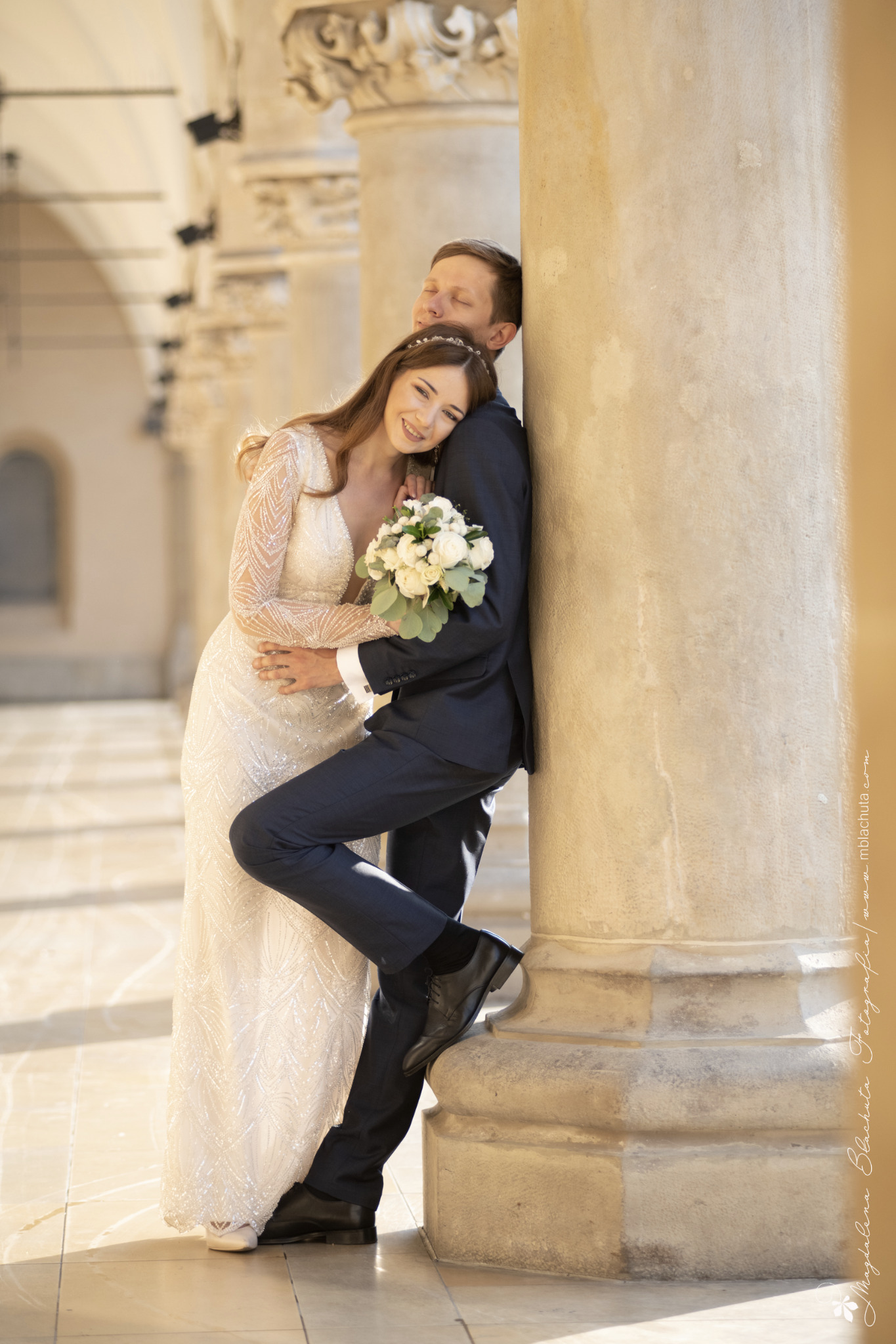 Para młoda w objęciach, oparta o kolumnę w słoneczny dzień. Panna młoda w sukni z długim rękawem, trzyma bukiet białych róż. Pan młody w granatowym garniturze.