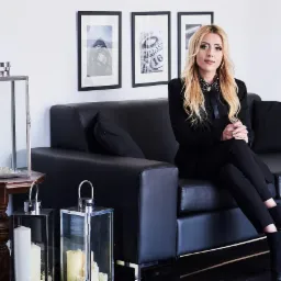 Kobieta w czarnym garniturze siedzi na czarnej skórzanej sofie w minimalistycznym wnętrzu, z metalowymi lampionami i czarno-białymi obrazami na ścianie.