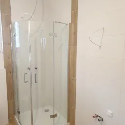 Narożna kabina prysznicowa z przezroczystego szkła, zamontowana na białym brodziku, na podłodze płytki imitujące drewno, białe płytki ścienne z pionowymi wstawkami imitującymi drewno, widoczne...