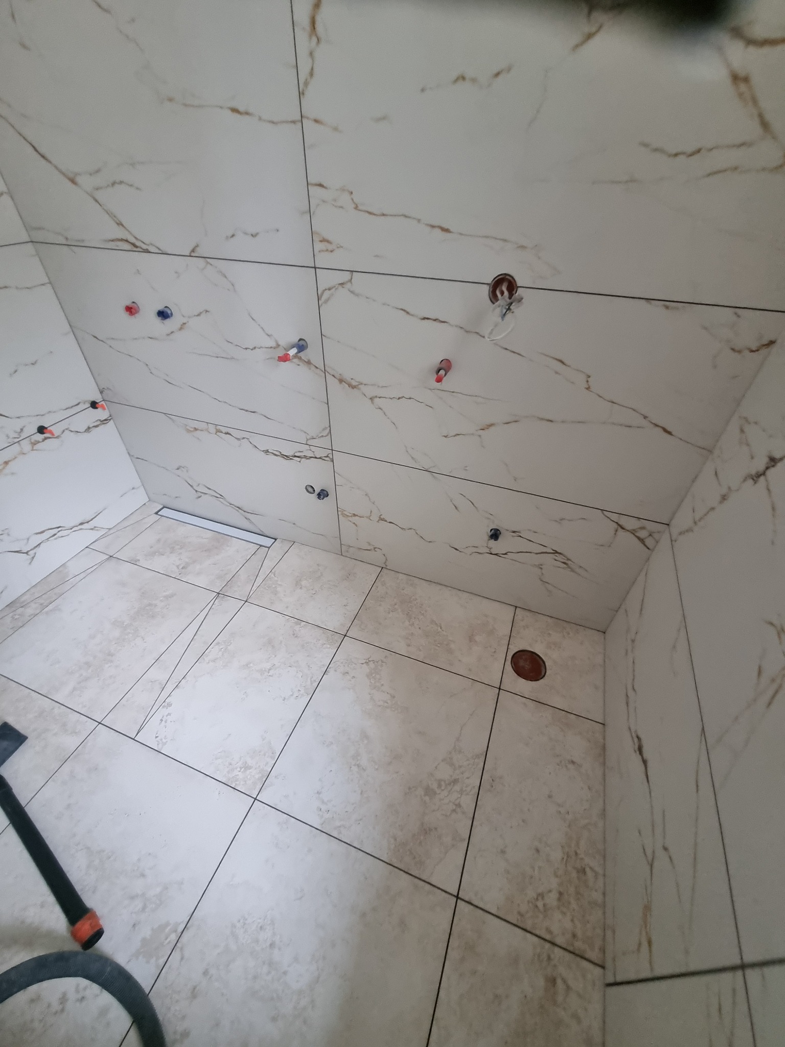 Wnętrze łazienki w trakcie remontu, widoczne płytki imitujące marmur na ścianach i podłodze, przygotowane otwory pod armaturę, odpływ liniowy w podłodze, fragment narzędzia glazurniczego.