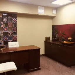 Wnętrze gabinetu z biurkiem, krzesłami, szafką z dekoracją zen (Buddha, bambus, świeca) i ścianą z dekoracyjnych szklanych płytek.