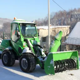 Kompaktowa, zielona maszyna AVANT z pługiem i frezarką do śniegu stoi na odśnieżonym placu, w tle budynki pokryte śniegiem i zalesione wzgórze w zimowej scenerii.