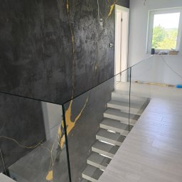 Team Glass - Szklana balustrada schodowa z czarną ścianą zdobioną złotym wzorem żyłkowania. Nowoczesne wykończenie wnętrza, jasne schody i podłoga.