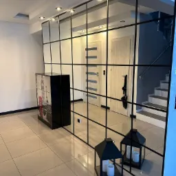 Nowoczesna szklana ścianka działowa z czarnymi profilami, dzieląca przestrzeń w jasnym wnętrzu. Widoczne drzwi i schody w tle, lampiony i szafka z motywem Londynu.