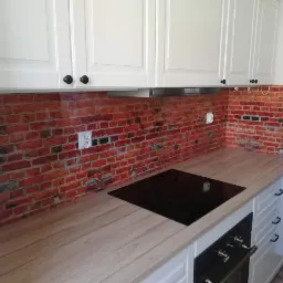 Szklana ściana kuchenna z motywem czerwonej cegły, zamontowana między blatem a białymi szafkami wiszącymi, z widocznym gniazdkiem elektrycznym.