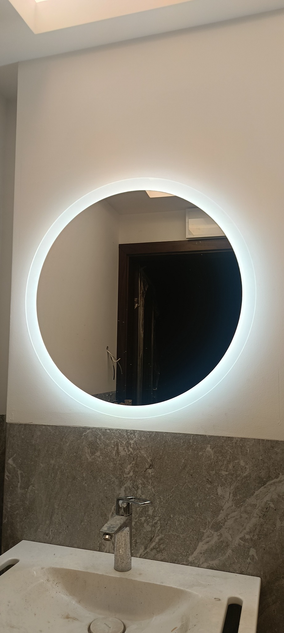 Okrągłe lustro z podświetleniem LED w łazience nad umywalką z szarą armaturą i marmurowym wykończeniem ściany. Widoczne odbicie wnętrza.