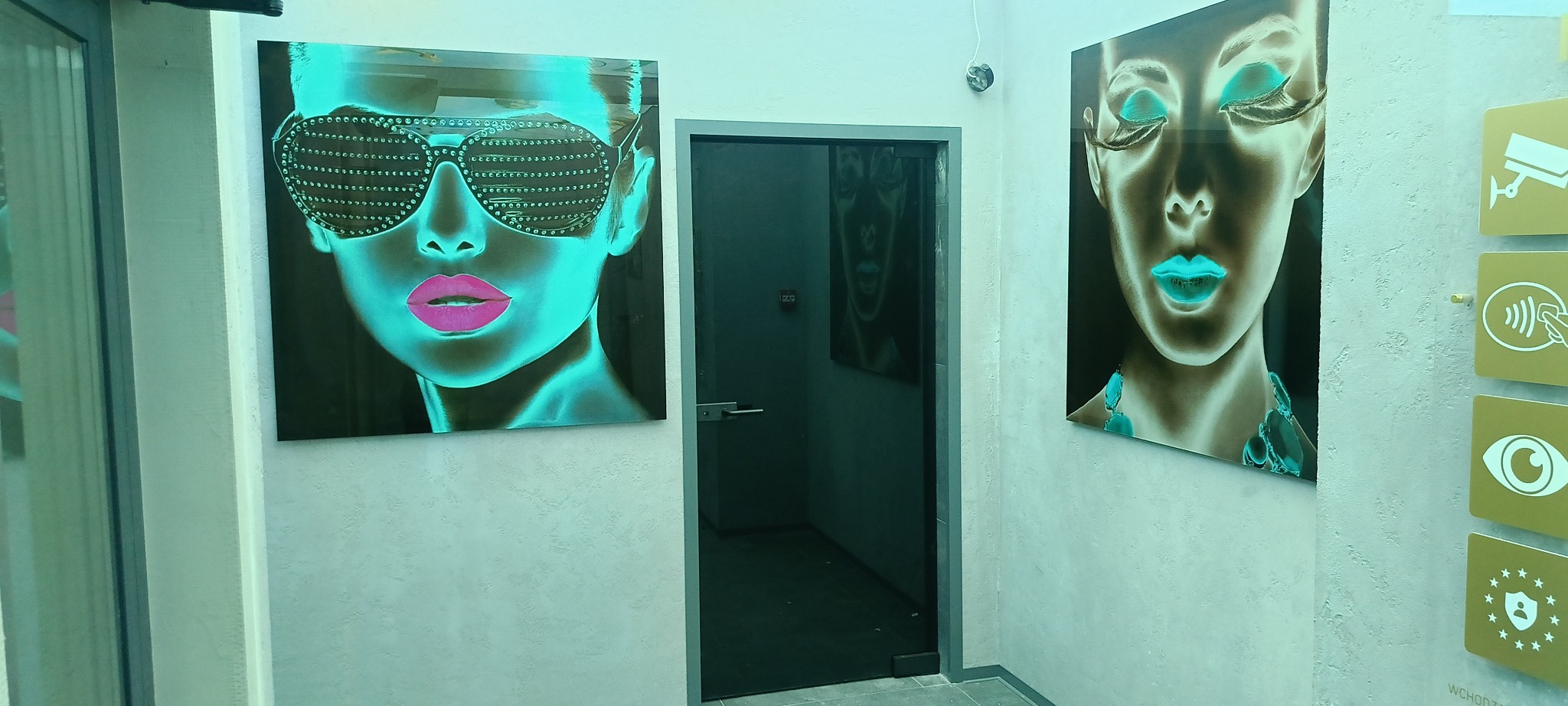 Szklane obrazy z motywem kobiecych twarzy w neonowych barwach, wiszące na ścianie w korytarzu. Widoczne drzwi i elementy systemu zabezpieczeń.