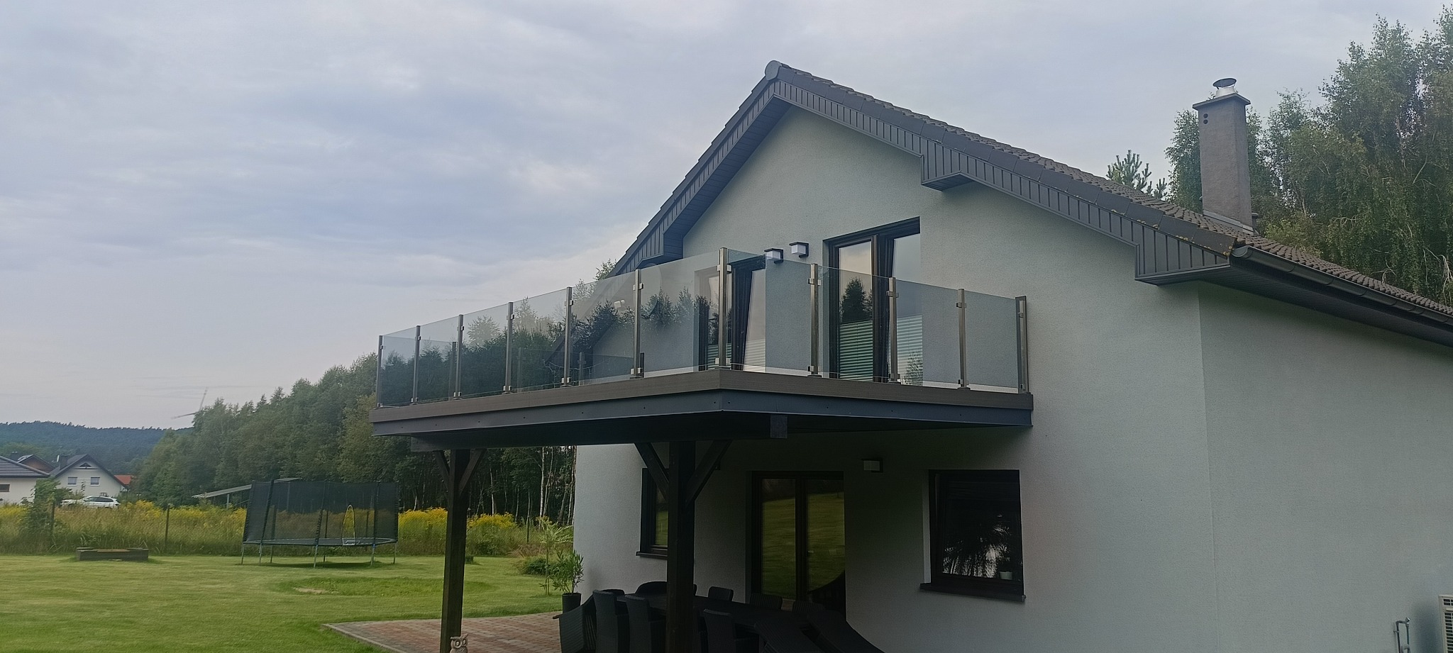 Nowoczesna, szklana balustrada balkonowa z metalowymi słupkami, zamontowana przy jasnym domu jednorodzinnym. Widok z dołu na tle zieleni.