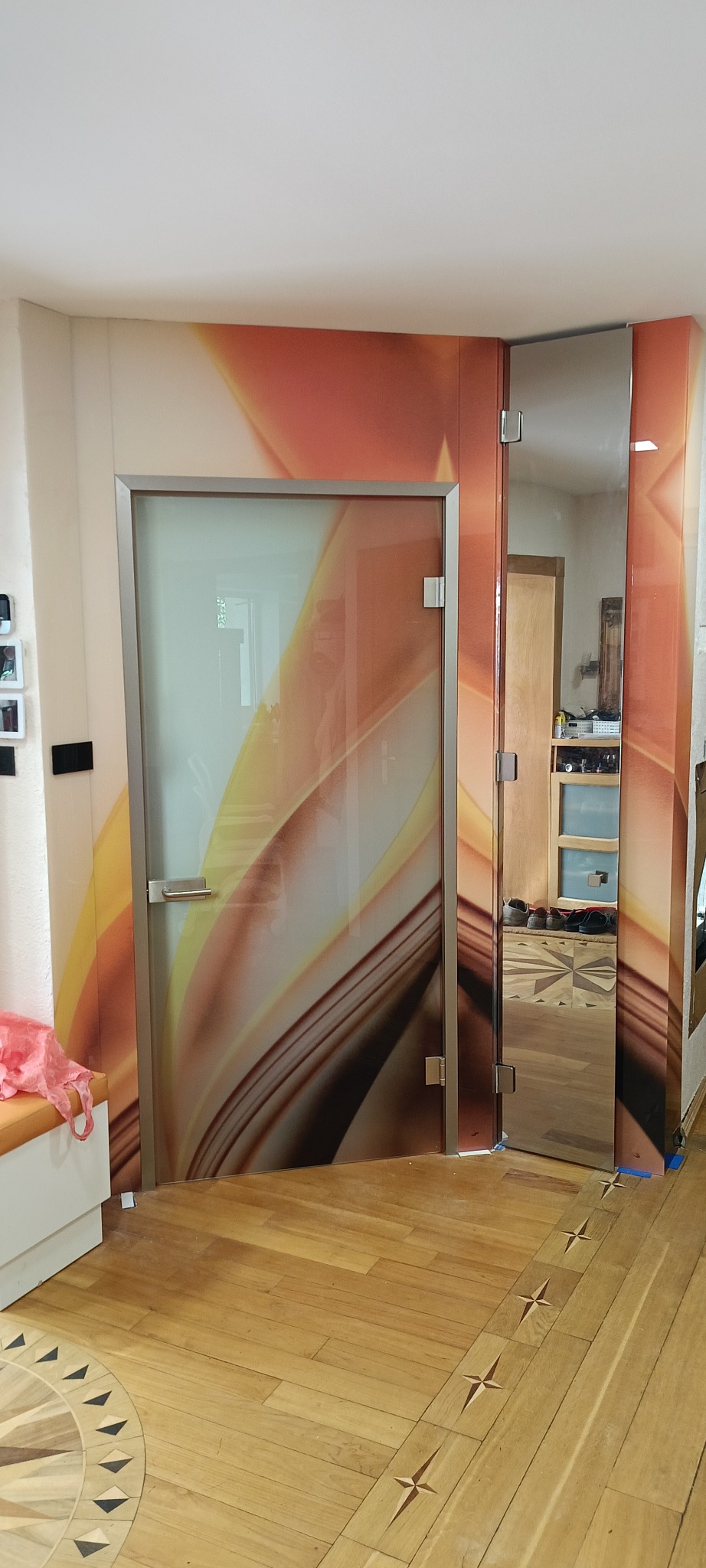 Szklane drzwi z abstrakcyjnym wzorem i lustrzana ściana w przedpokoju. Widoczna drewniana podłoga z dekoracyjnym wzorem. Całość tworzy nowoczesny i stylowy wystrój.