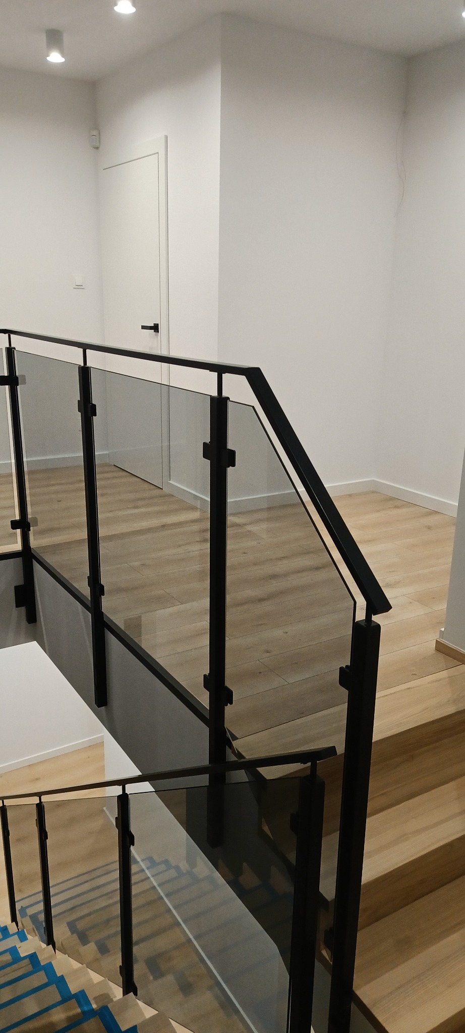 Nowoczesna, szklana balustrada schodowa z czarnymi elementami mocującymi. Widok z góry na schody z drewnianymi stopniami i zabezpieczeniem w postaci niebieskiej taśmy.