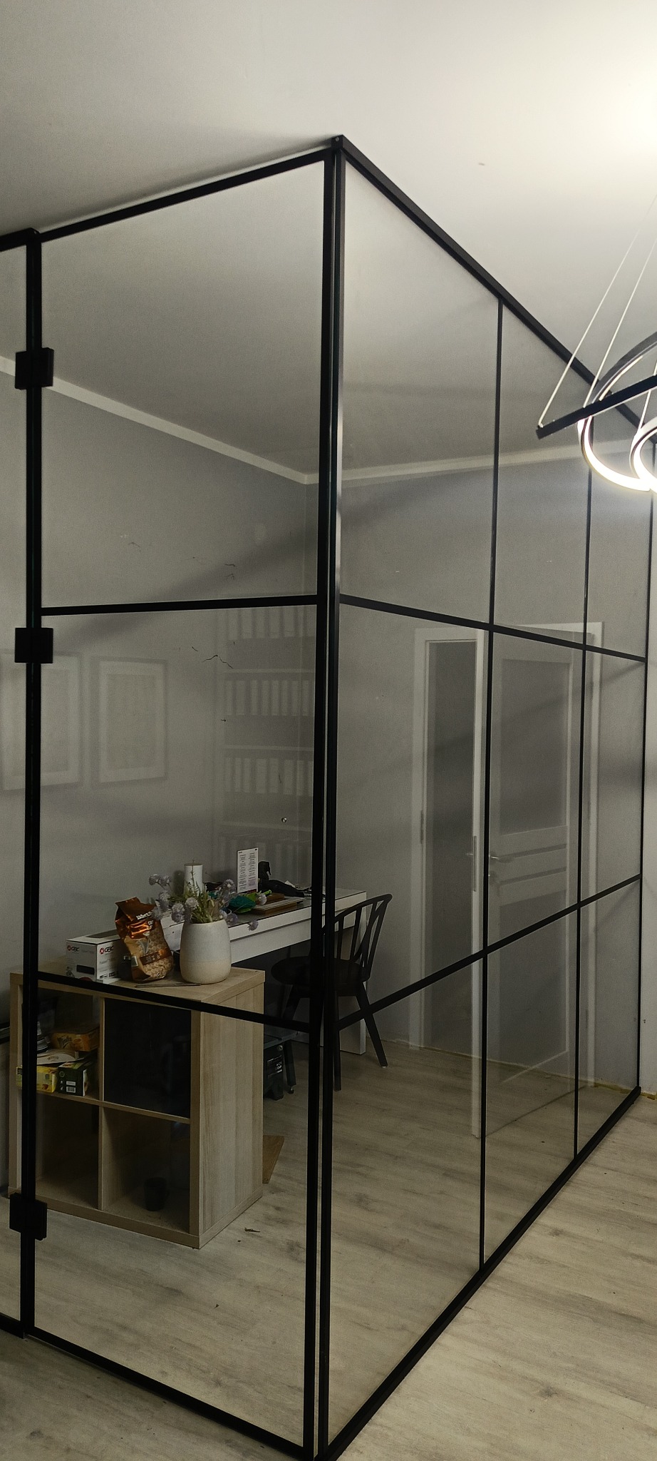 Nowoczesna szklana ścianka działowa z czarnymi profilami, oddzielająca przestrzeń biurową w jasnym wnętrzu. Widoczny fragment biurka i regału.