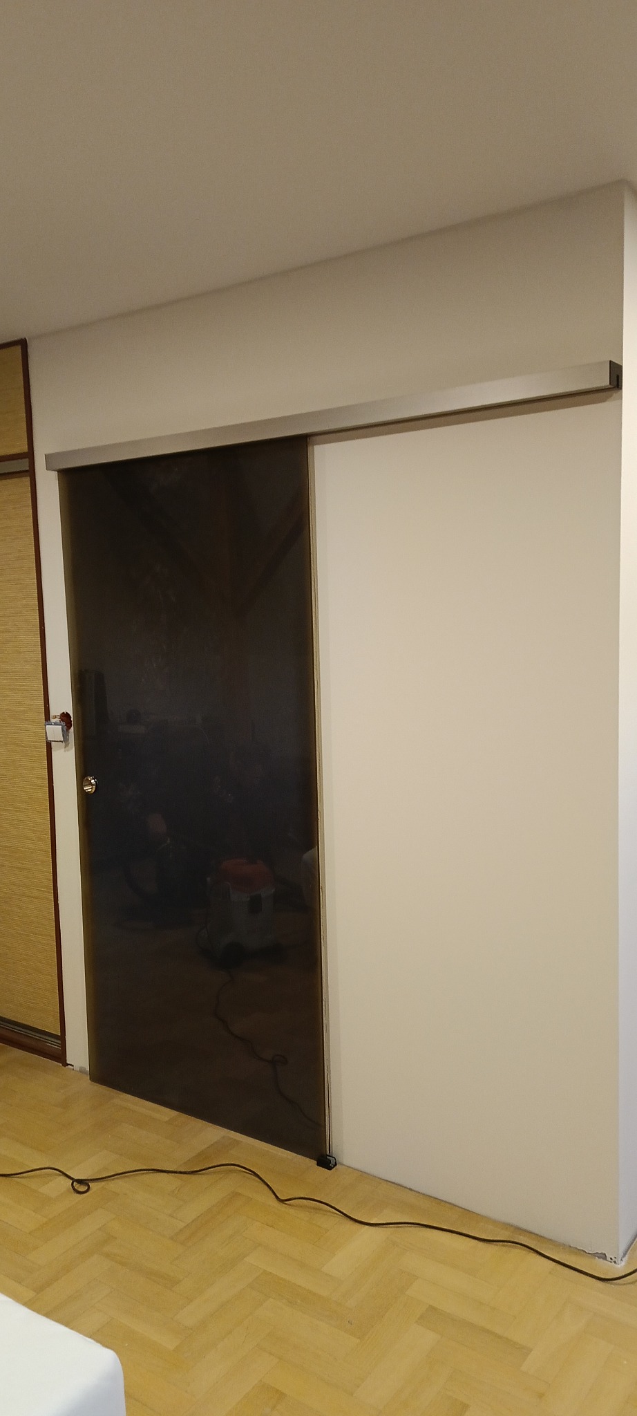 Szklane drzwi przesuwne w systemie szynowym, z widocznym odbiciem wnętrza. Minimalistyczny design, jasne ściany i parkiet w jodełkę. Detal wykończenia wnętrza.