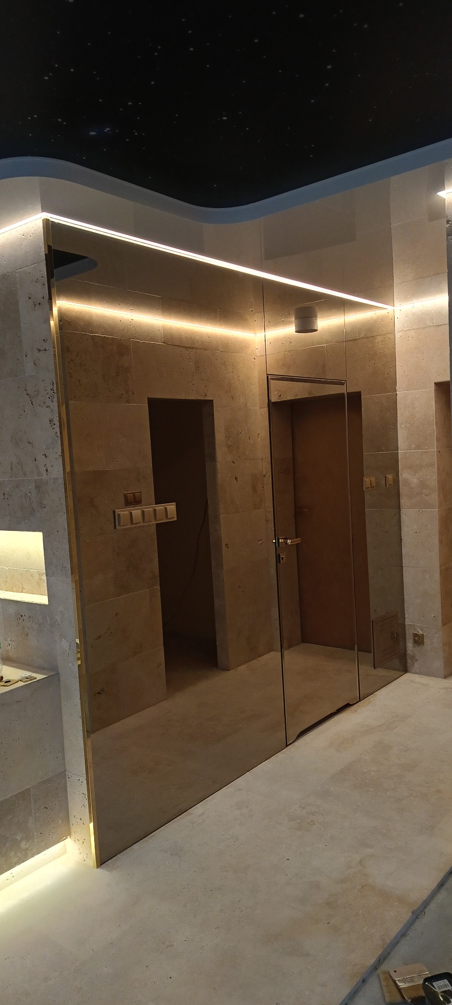 Nowoczesne lustro w łazience z podświetleniem LED, odbijające drzwi i fragment sufitu z motywem gwiazd. Wykończenie w odcieniach beżu i złota.