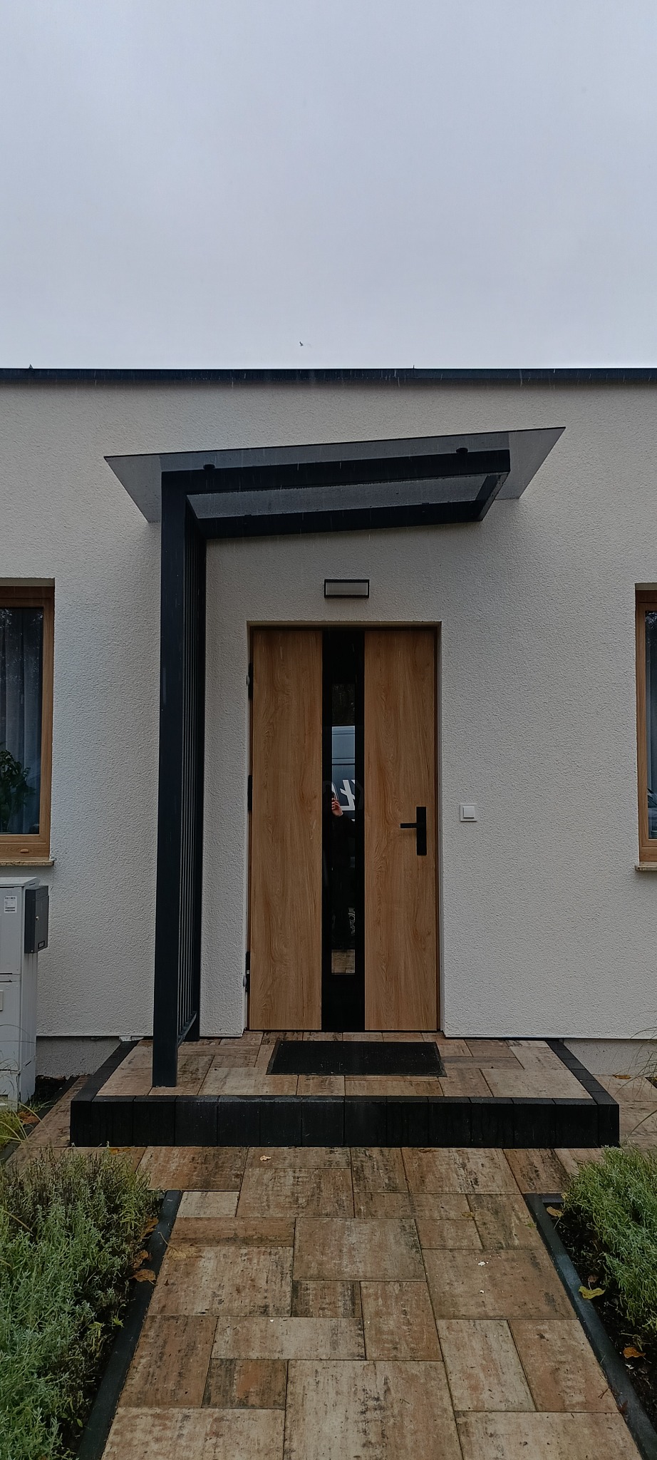 Nowoczesne wejście do domu z drewnianymi drzwiami i szklanym zadaszeniem wspartym na czarnej konstrukcji. Ułożona kostka brukowa prowadzi do drzwi.