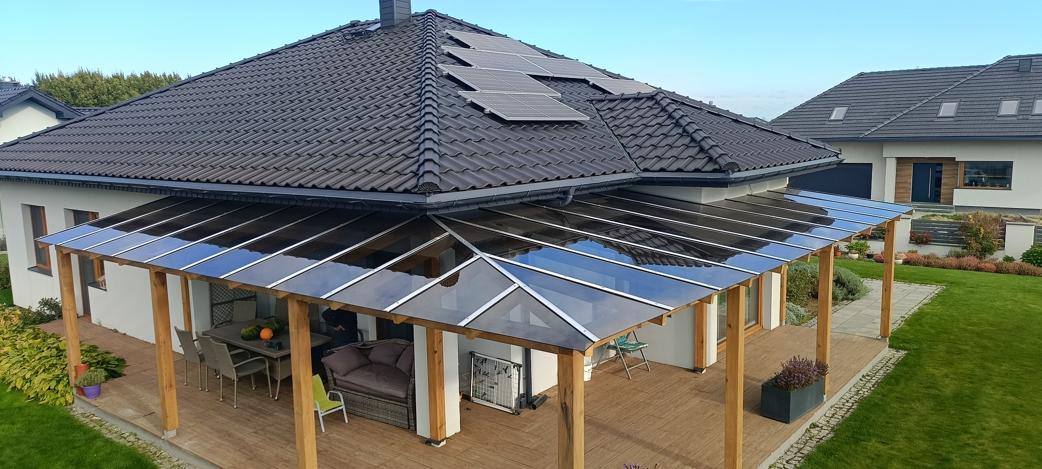 Zadaszenie tarasu z przezroczystymi panelami, wsparte na drewnianych słupach, przylegające do domu z panelami słonecznymi na dachu. Widok z góry na taras.