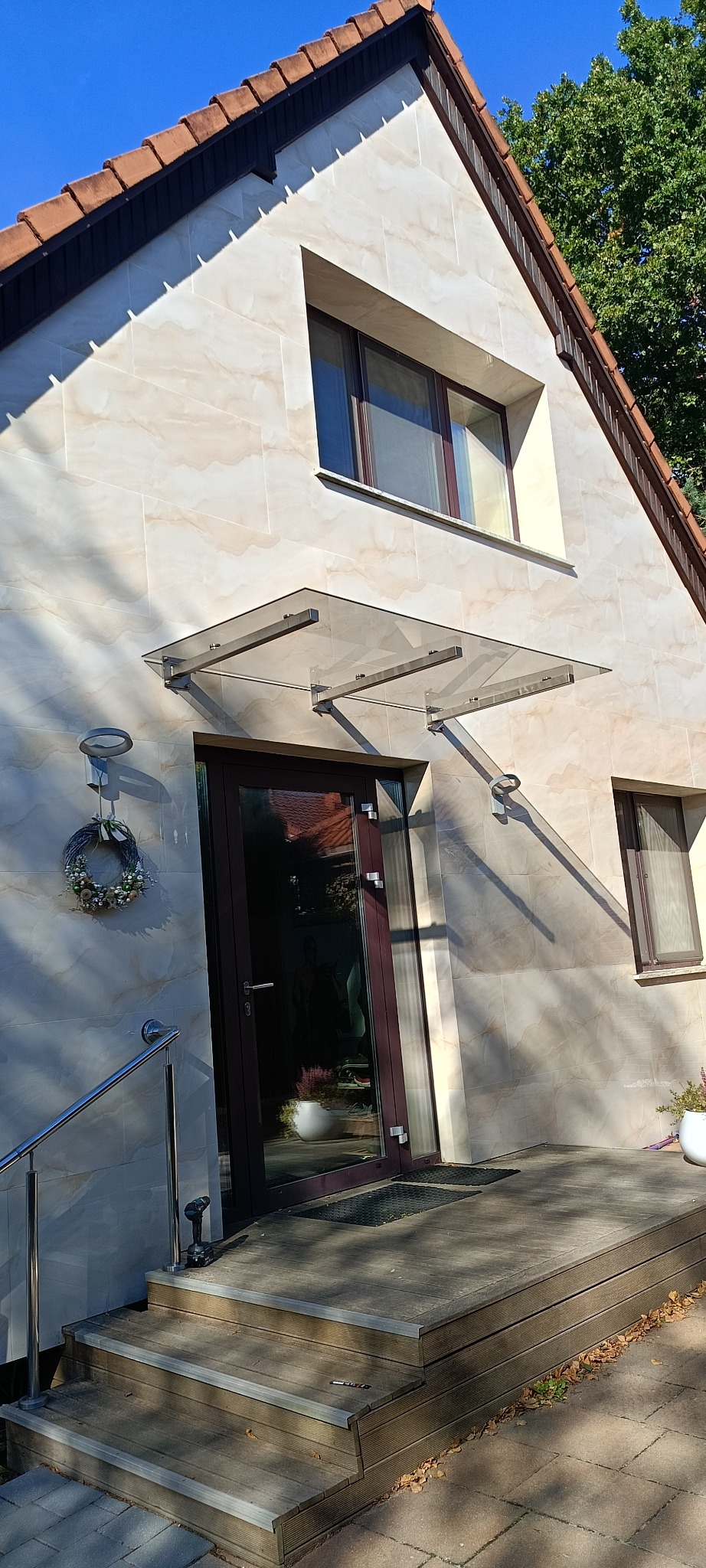 Dom z przeszklonym zadaszeniem nad wejściem, wspartym na metalowych wspornikach. W tle widoczne okno i fragment dachu. Na schodach leży wkrętarka.