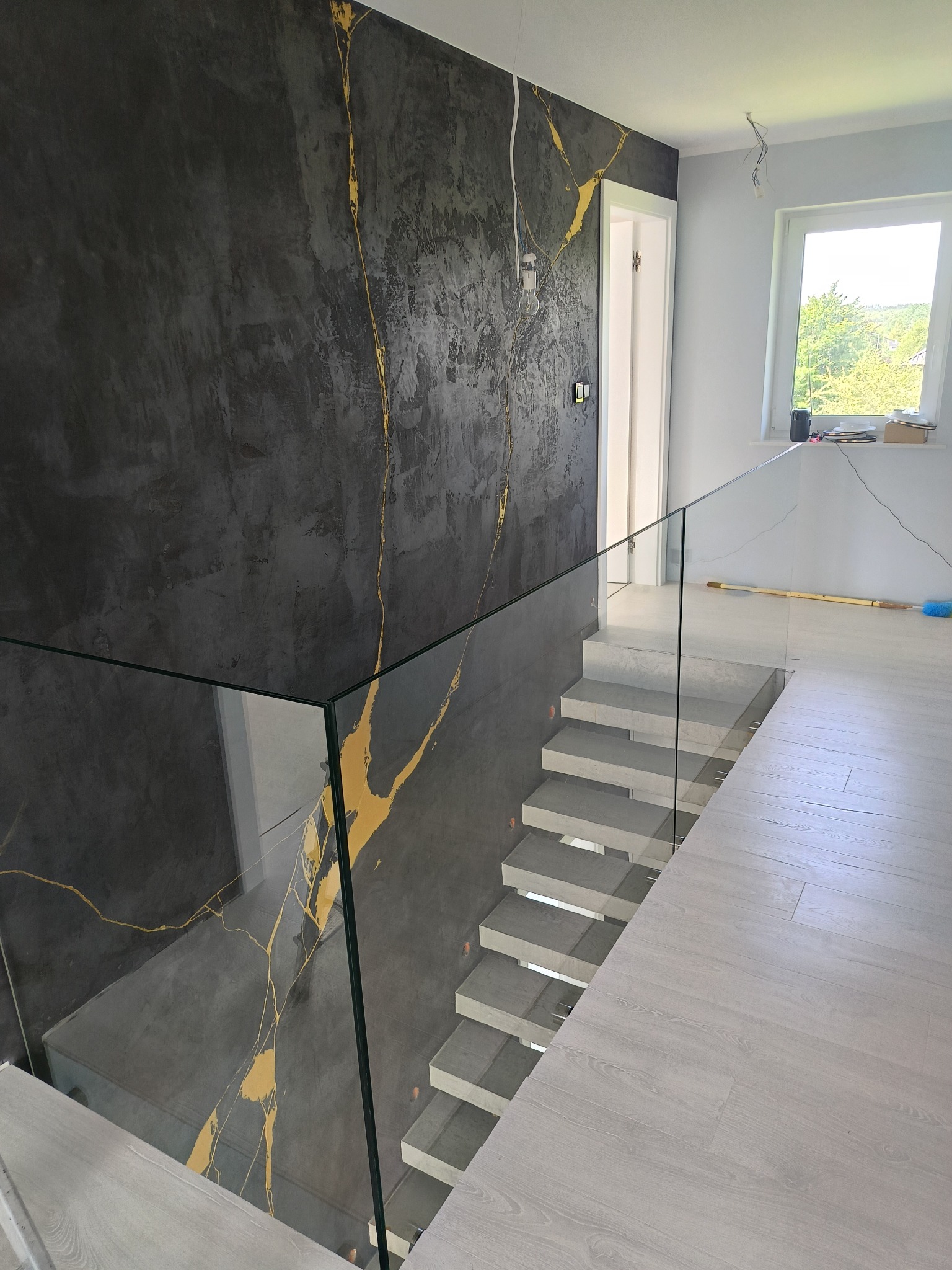 Szklana balustrada schodowa z czarną ścianą zdobioną złotym wzorem żyłkowania. Nowoczesne wykończenie wnętrza, jasne schody i podłoga.