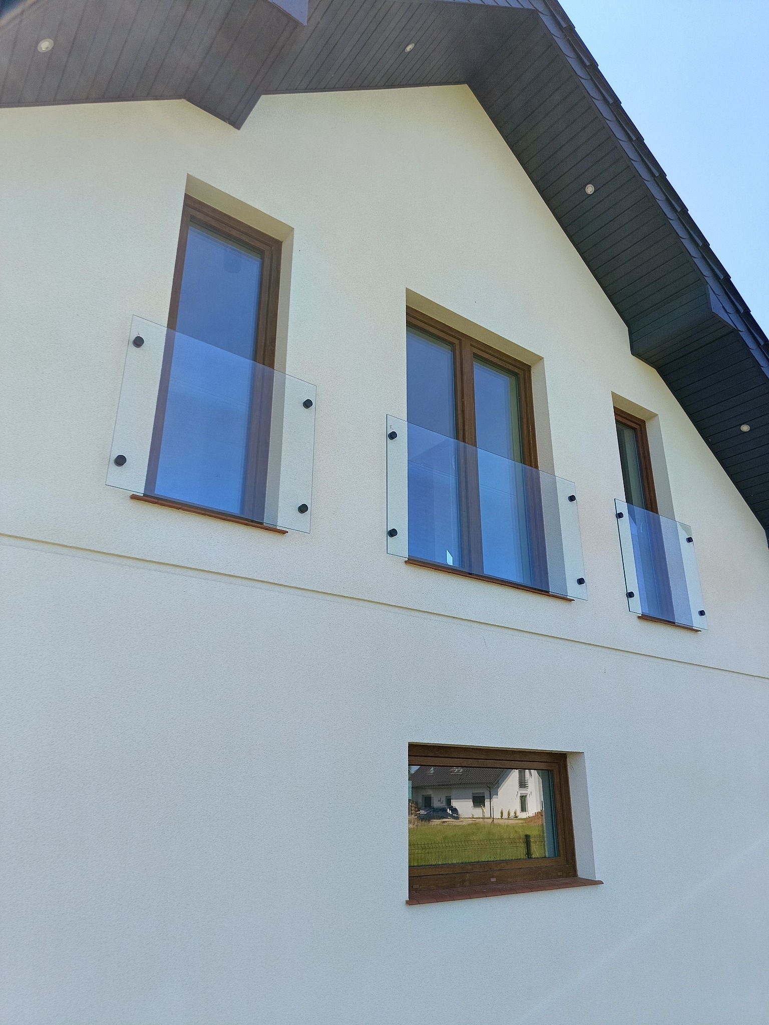 Elewacja domu z trzema oknami na piętrze, zabezpieczonymi szklanymi balustradami mocowanymi na cztery punkty. W dole okno z odbiciem budynku.