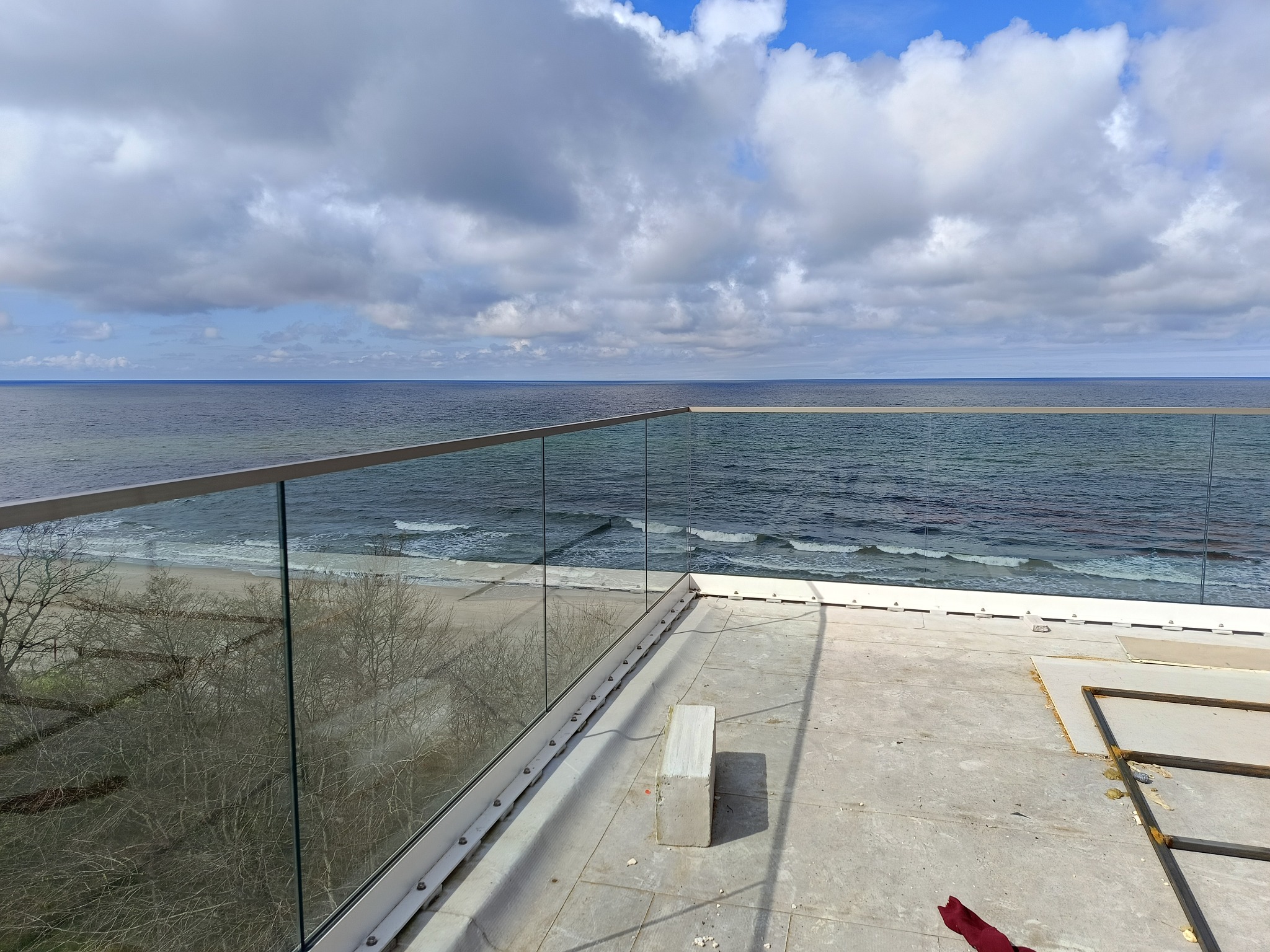 Szklana balustrada balkonowa z widokiem na morze w słoneczny dzień. Minimalistyczny design, srebrne okucia, błękitne niebo z chmurami odbijające się w tafli szkła.