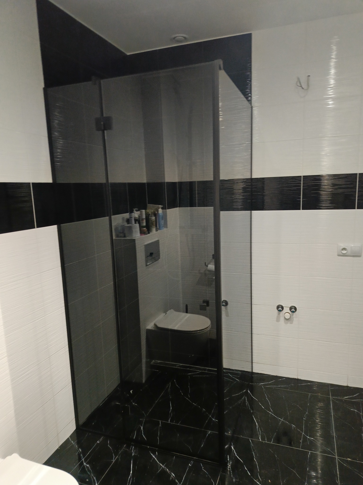 Nowoczesna kabina prysznicowa ze szkła hartowanego w łazience z czarno-białymi płytkami. Widoczne detale wykończenia i armatura. Marmurowa podłoga.