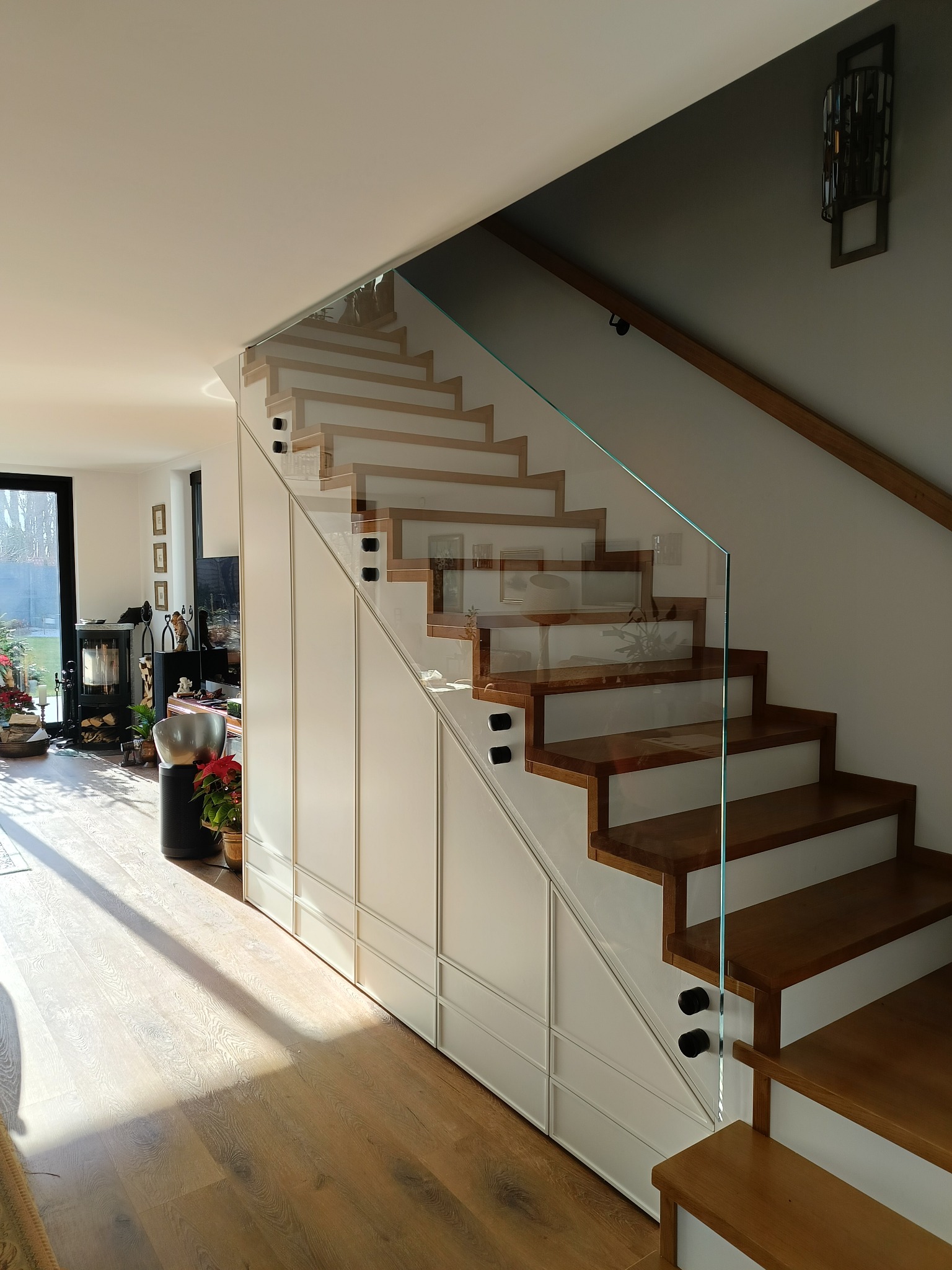Nowoczesne schody z drewnianymi stopniami i szklaną balustradą, wkomponowane w zabudowę meblową. Jasne wnętrze z widokiem na ogród.